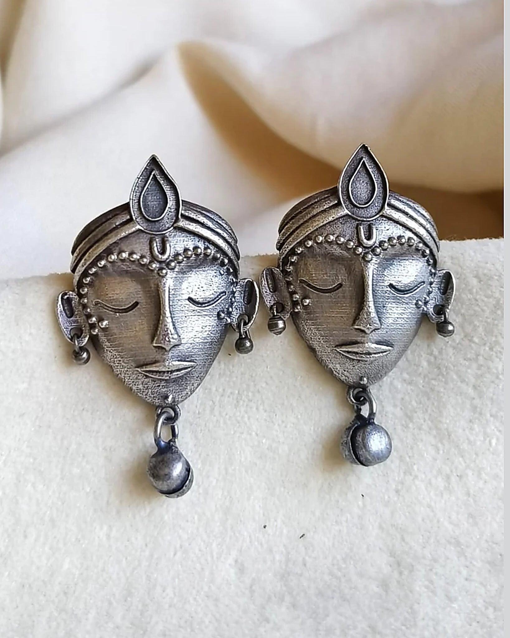 Oxidised silver-look brass stud earrings shaped as crowned goddess faces, each with a small bell drop