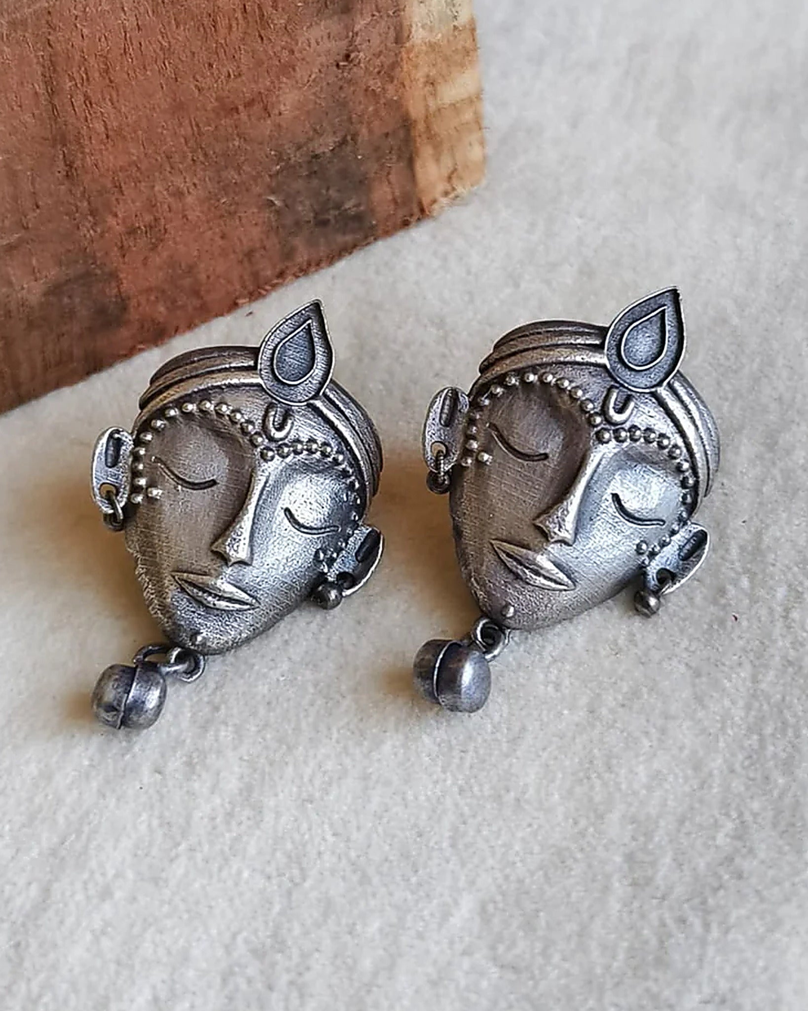 Oxidised silver-look brass stud earrings shaped as crowned goddess faces, each with a small bell drop