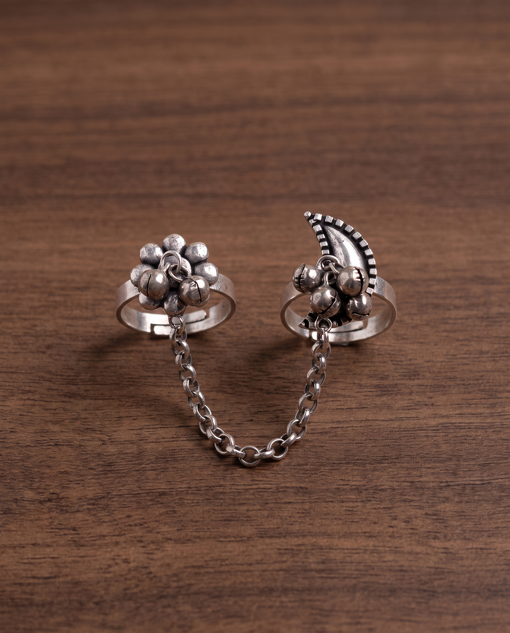 Close-up of the Pushkara two-finger ring on dark walnut wood, showing the rollo chain links and hanging ghungroo bells