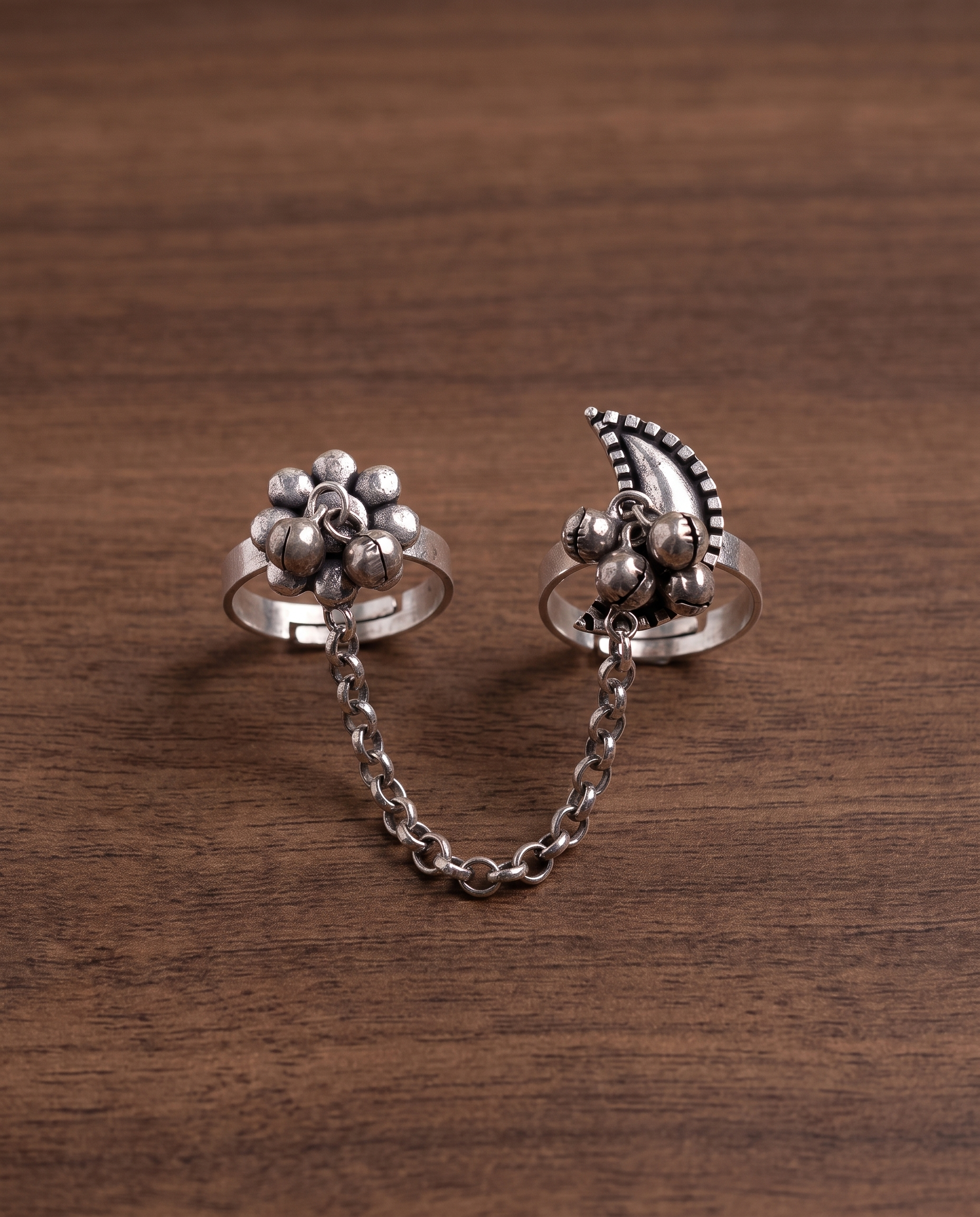 Close-up of the Pushkara two-finger ring on dark walnut wood, showing the rollo chain links and hanging ghungroo bells