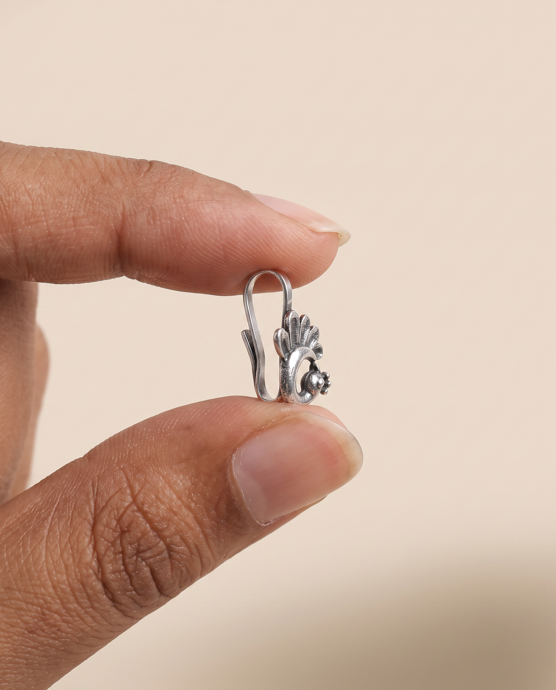 Morni peacock nose pin held between fingertips in profile, showing its small size and the flat U-shaped clip that needs no piercing