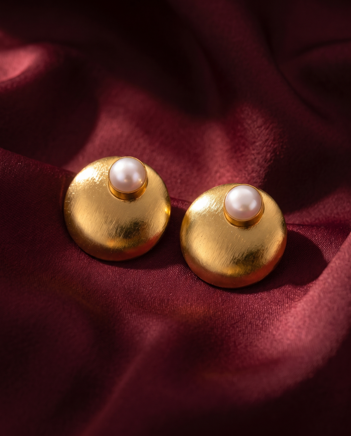 Gold-tone anti-tarnish brass stud earrings each set with a domed freshwater pearl, styled on burgundy silk