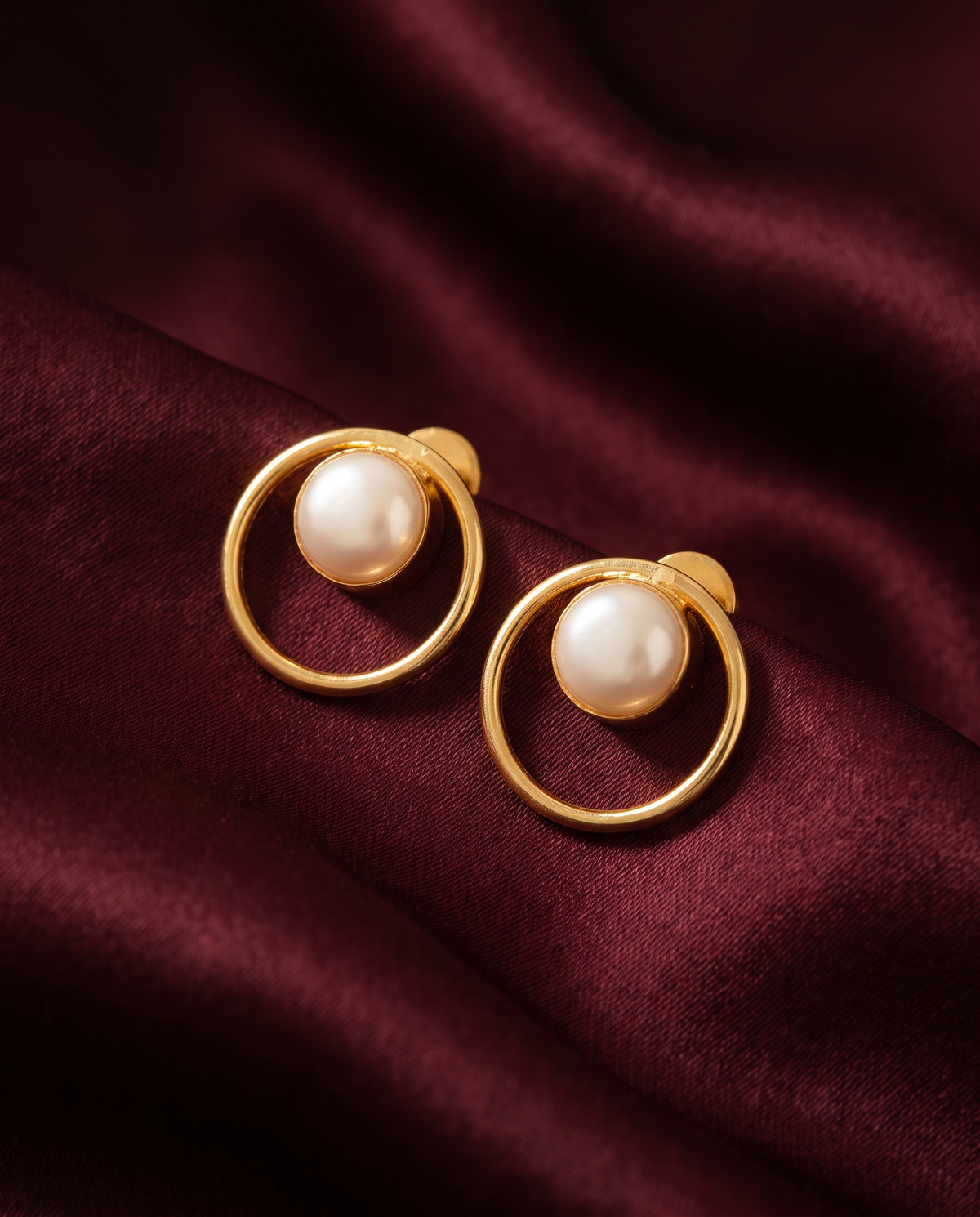 Gold-tone anti-tarnish brass stud earrings with a single pearl inside a round frame, styled on burgundy silk
